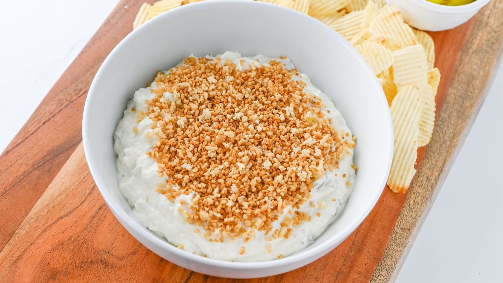A bowl of creamy dip topped with crumbled breadcrumbs sits on a wooden board, surrounded by wavy potato chips.
