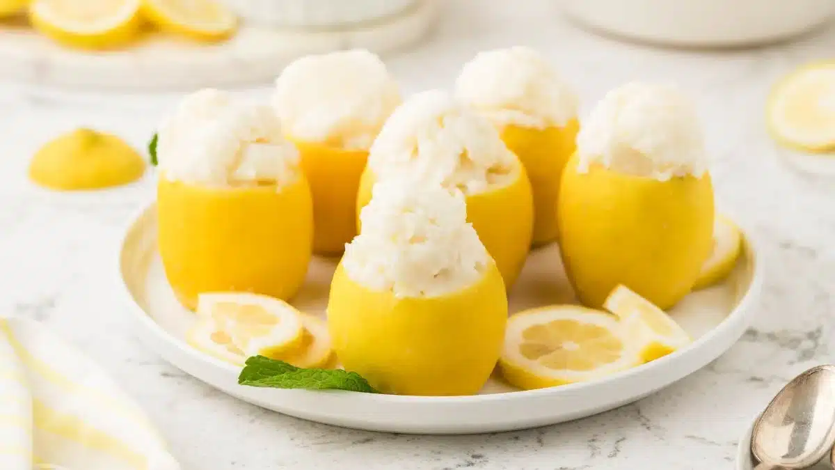 A plate of hollowed-out lemons filled with a scoop of lemon sorbet, garnished with fresh lemon slices and a sprig of mint on a white surface.