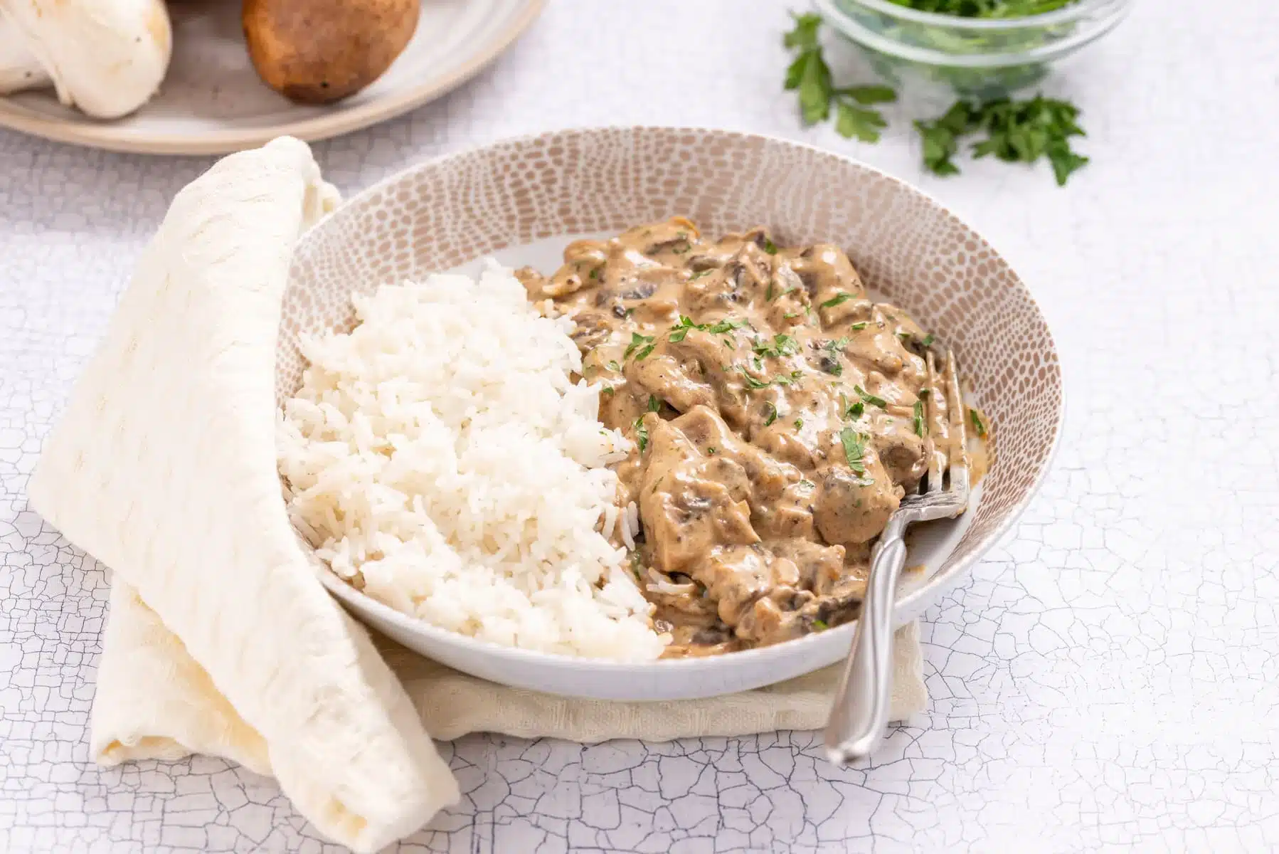 A bowl of white rice and creamy beef stroganoff garnished with chopped parsley, served with a fork and a napkin on the side. A plate with mushrooms and a bowl of herbs are in the background.