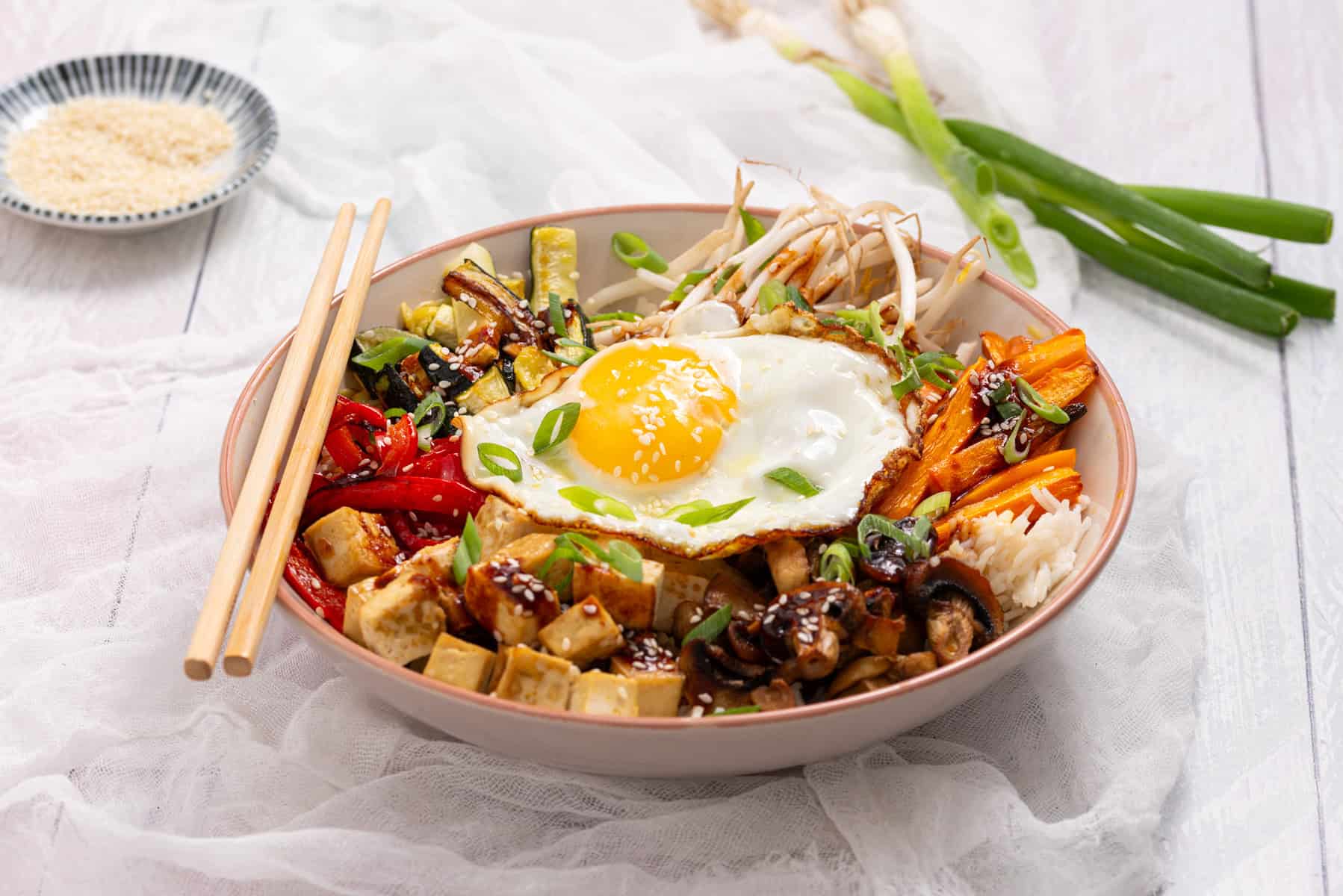 A bowl of bibimbap with a sunny-side-up egg, tofu, sautéed vegetables, shredded carrots, bean sprouts, and rice, garnished with green onions, with chopsticks resting on the bowl.