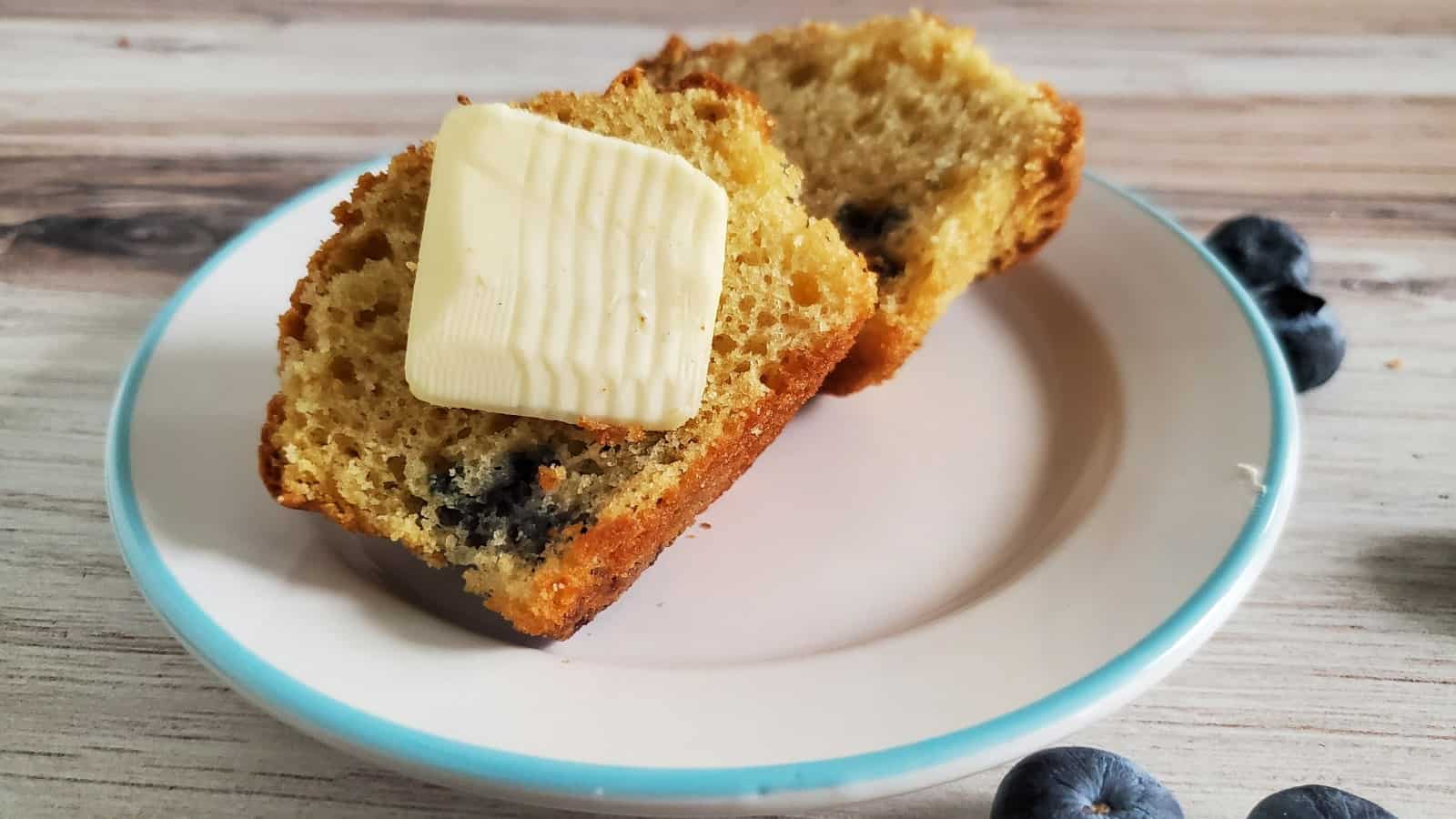 Two slices of blueberry bread on a white plate with a blue rim; one slice is topped with a pat of butter. A few fresh blueberries are beside the plate on a wooden surface.