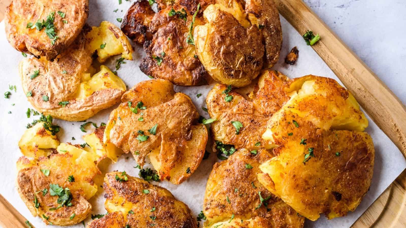 Golden, crispy smashed potatoes garnished with chopped herbs are arranged on parchment paper atop a wooden board. The potatoes are browned and slightly broken open, revealing their soft insides.