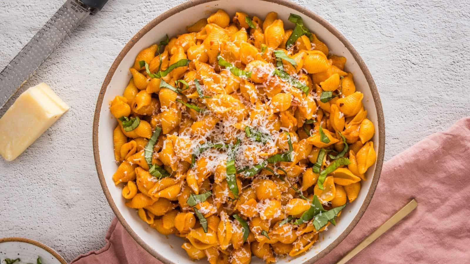 A bowl of shell pasta in a creamy orange sauce, topped with grated cheese and fresh basil, sits on a textured white surface next to a block of cheese and a pink napkin.