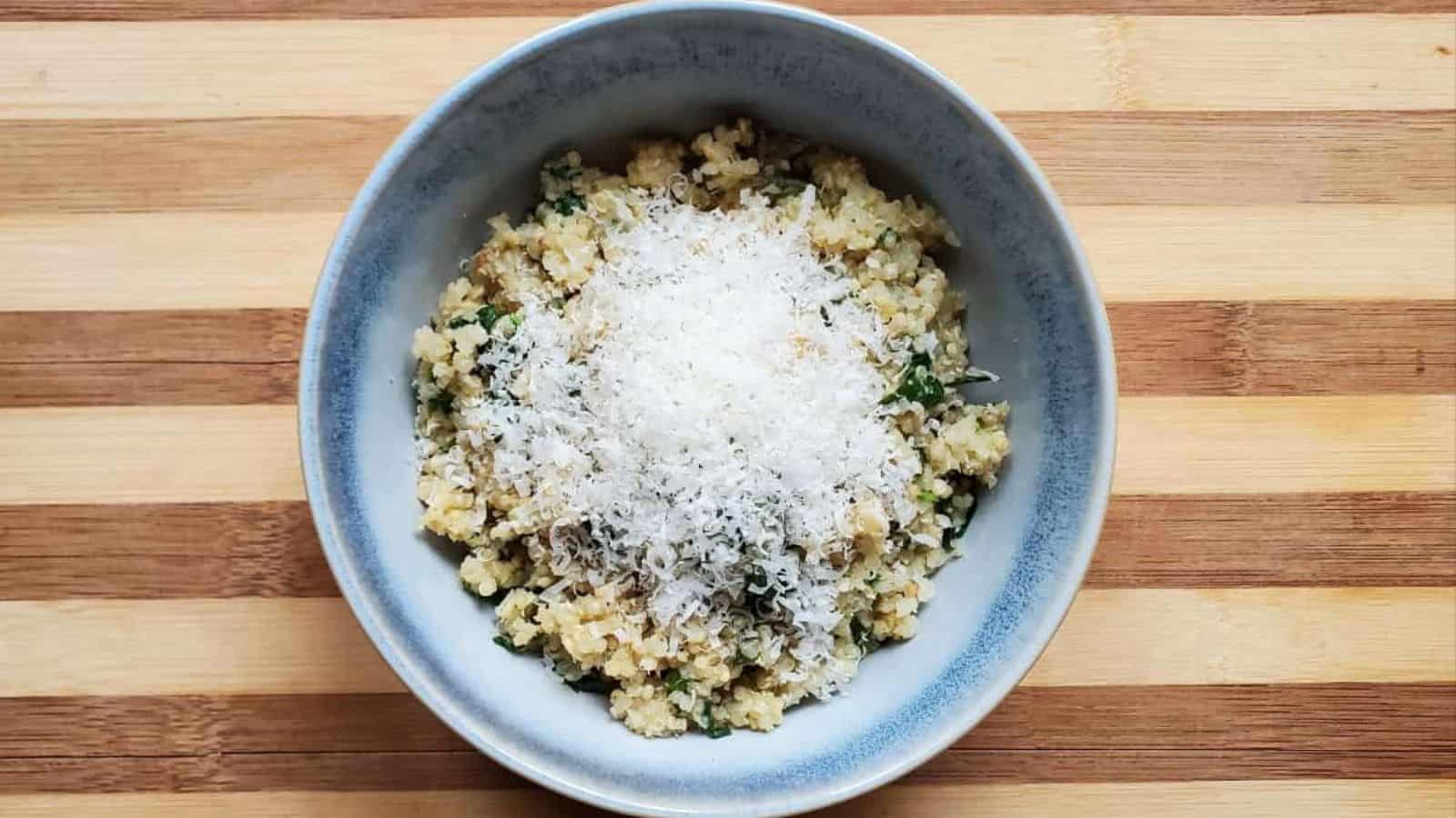 A blue-gray bowl filled with quinoa mixed with greens, topped with a generous layer of grated cheese, sits on a striped wooden surface.