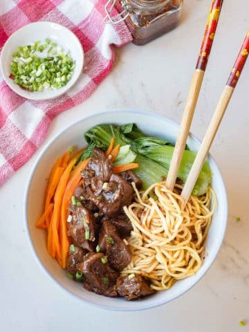 A bowl of noodles topped with braised beef, bok choy, and carrots. Chopsticks rest on the bowl. A jar of sauce and a small bowl of chopped green onions sit nearby on a white surface with a red checkered cloth.