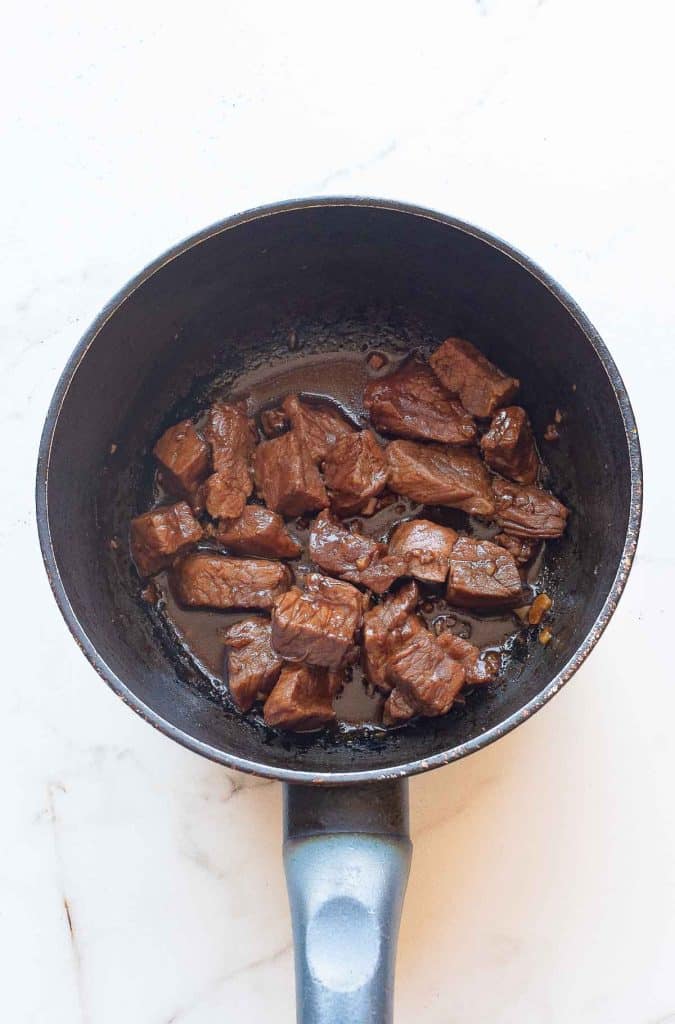 A black saucepan containing browned chunks of beef sits on a white marble surface. The beef pieces are cooked and coated with a glossy sauce.