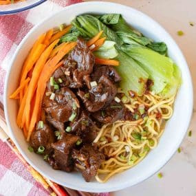 A bowl of noodle soup topped with braised beef, sliced carrots, bok choy, and chopped green onions, placed on a red and white checkered cloth. Chopsticks are beside the bowl.