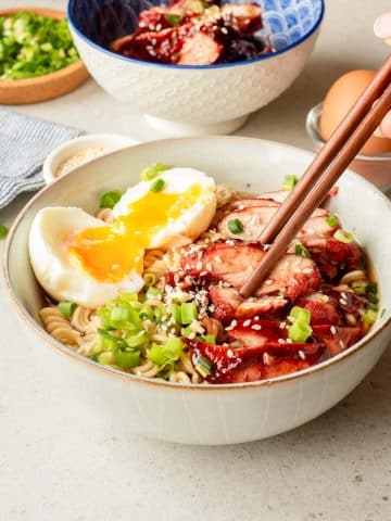 A bowl of ramen topped with sliced char siu pork, a soft-boiled egg with a runny yolk, sliced green onions, and sesame seeds, with chopsticks picking up some noodles. Another bowl and toppings are in the background.
