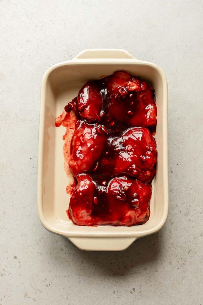 Four pieces of raw chicken coated in a dark red sauce are arranged in a rectangular beige baking dish on a light, speckled countertop.