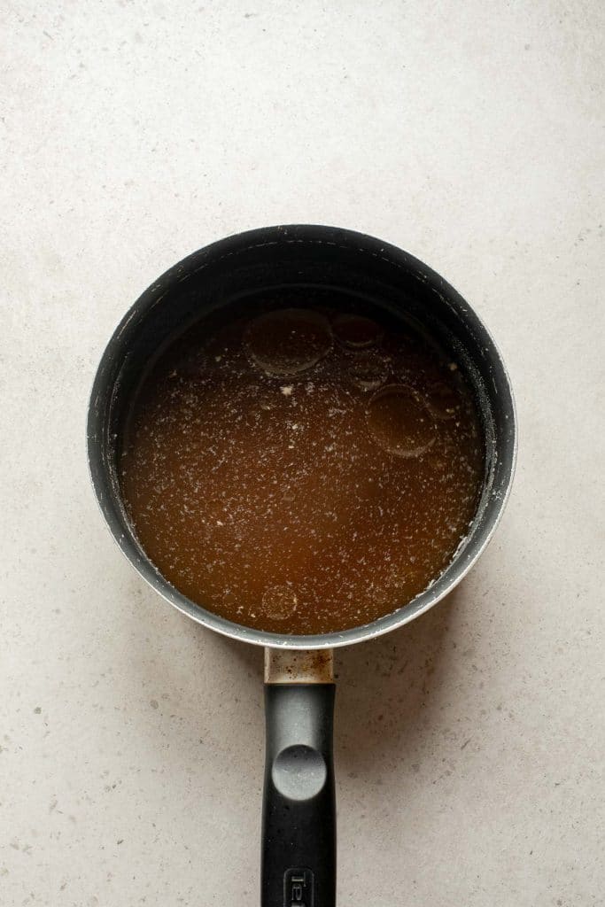 A small black saucepan with a handle contains brown liquid with visible oil droplets on the surface, set on a light-colored countertop.