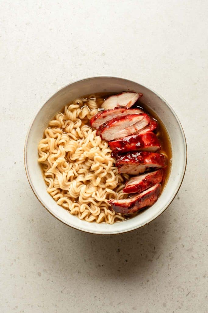 A bowl of ramen noodles in broth, topped with several slices of glazed, roasted chicken, set on a light-colored surface.
