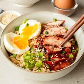 A bowl of ramen noodles topped with sliced roast pork, a soft-boiled egg with a runny yolk, chopped green onions, and sesame seeds, with chopsticks resting on the bowl.