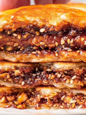 Close-up of three flaky pastries stacked on a plate, with a visible filling of nuts and sesame seeds in a caramelized sauce. A hand is holding the top pastry, showing its textured, gooey interior.