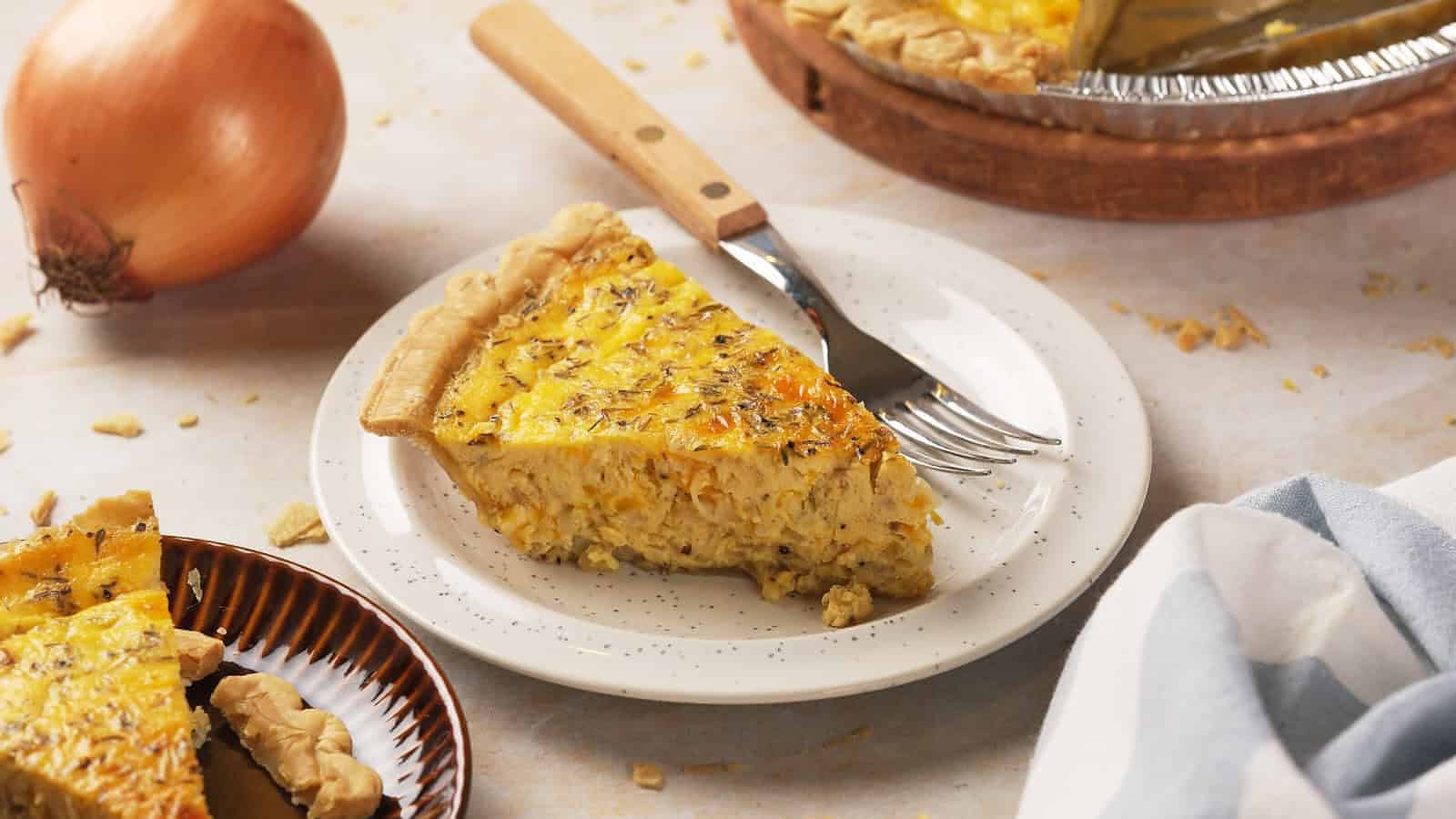 A slice of quiche with a golden, flaky crust sits on a white plate with a fork and knife. An onion and a partially sliced quiche are in the background on a light-colored surface.