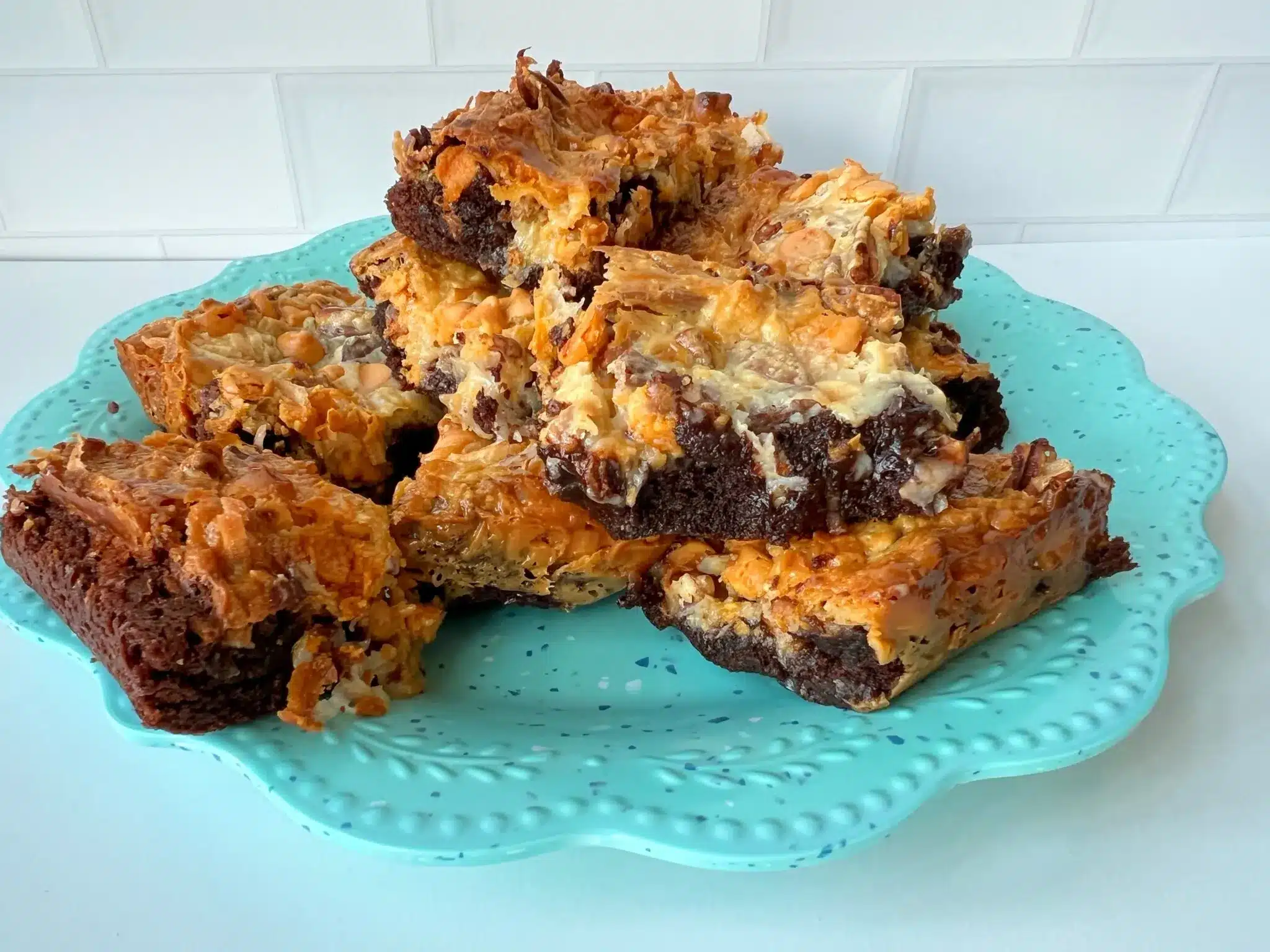 A turquoise plate holds several gooey chocolate and butterscotch bars with a marbled, textured topping, set against a white tile background.