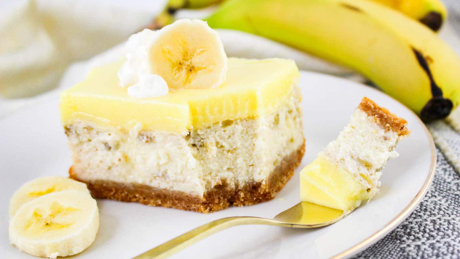 A slice of banana cream cheesecake with a graham cracker crust, topped with banana slices and whipped cream, is served on a plate with a fork—perfect for fans of classic cheesecake or banana pudding treats. Whole bananas sit in the background.