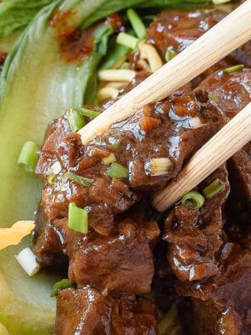 A close-up of chopsticks picking up pieces of braised beef over noodles, with sliced carrots and leafy bok choy in a bowl. The dish is garnished with green onions.
