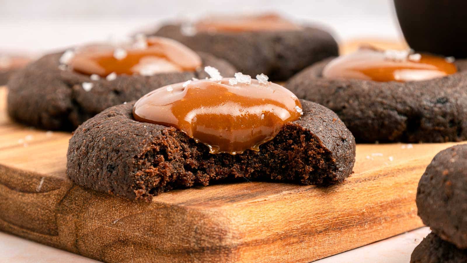 A close-up of chocolate cookies topped with caramel and sea salt, with one cookie showing a bite taken out, displayed on a wooden board.