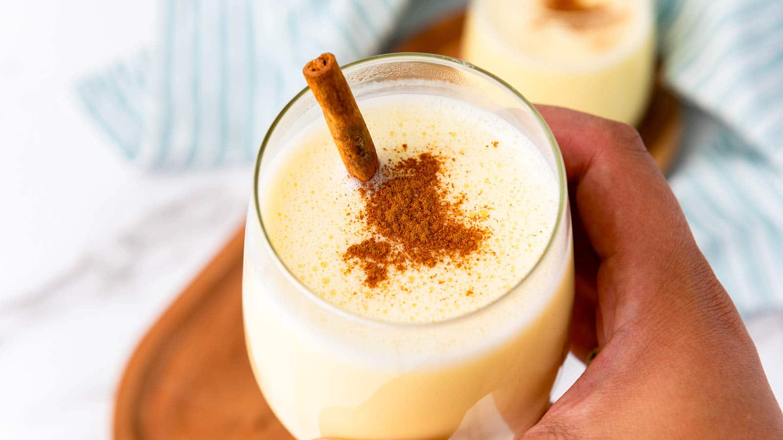 A hand holds a glass of creamy yellow homemade eggnog topped with ground cinnamon and a cinnamon stick, with a striped cloth in the background.