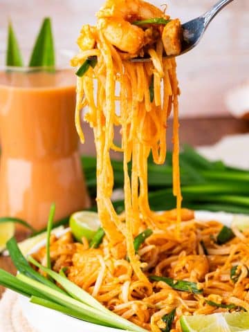 A close-up of a plate of Pad Thai noodles with shrimp, garnished with green onions and lime wedges. A fork lifts a serving of noodles above the plate. A glass of orange-colored drink is in the background.