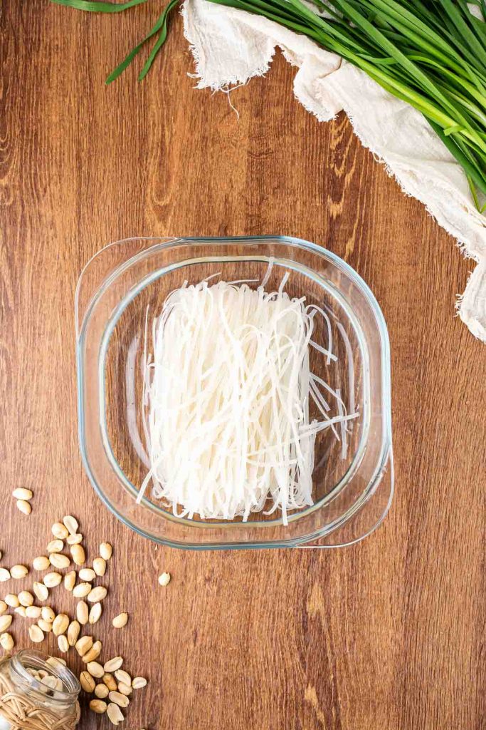 A glass bowl filled with uncooked rice noodles sits on a wooden table, surrounded by raw peanuts, twine, green herbs, and a white cloth napkin.