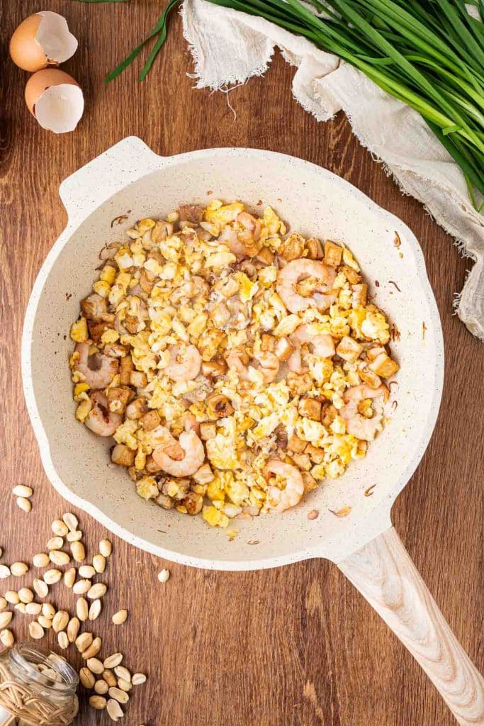 A white frying pan on a wooden table contains scrambled eggs, shrimp, tofu cubes, and chopped vegetables. Scallions, peanuts, eggshells, and a cloth napkin are nearby as food props.