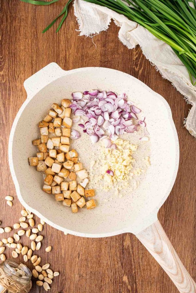 A white skillet on a wooden surface holds diced tofu, chopped red onion, and minced garlic in separate sections. Fresh chives, peanuts, and a garlic bulb are nearby on the table.