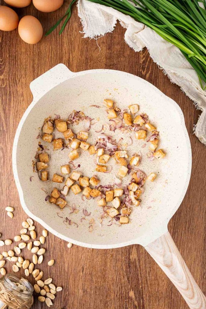 A white skillet on a wooden surface contains sautéed diced tofu and sliced red onions. Surrounding the skillet are eggs, peanuts, green onions, garlic, and a linen cloth.