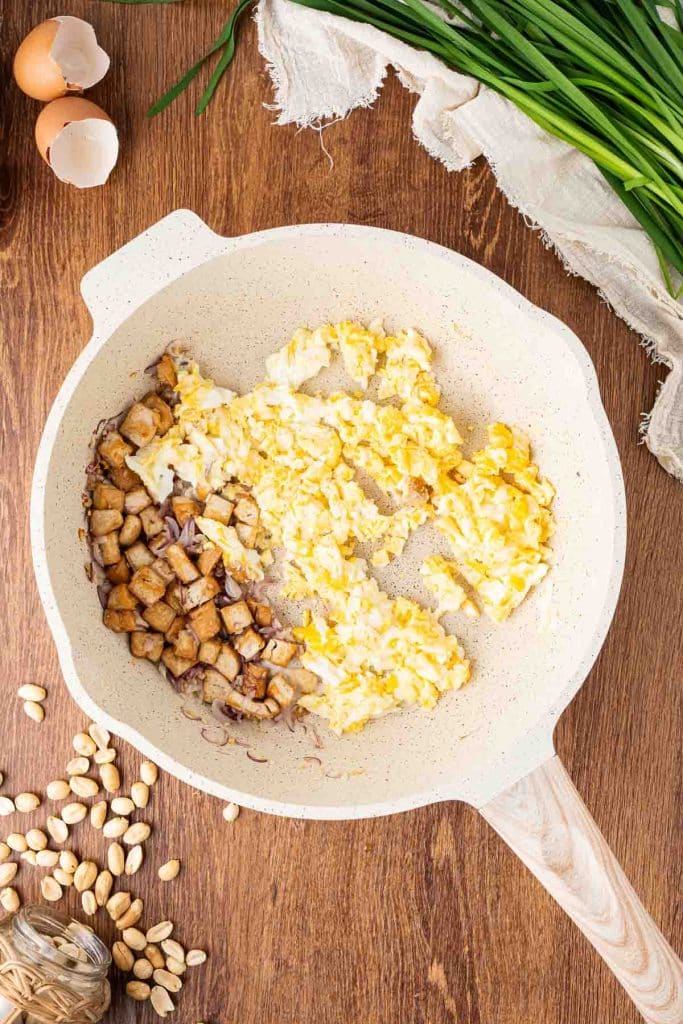 A light-colored skillet on a wooden surface contains scrambled eggs and browned tofu cubes. Scallions, peanuts, garlic, and open eggshells are scattered nearby, with a beige cloth at the top.