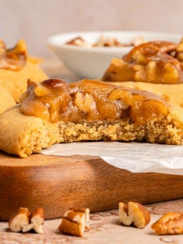 A close-up of pecan cookies topped with caramel and chopped pecans on parchment paper, with one cookie partially eaten, resting on a wooden board. Scattered pecan pieces and a bowl of pecans are in the background.
