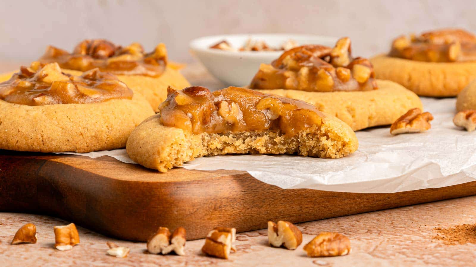 Close-up of pecan-topped cookies on a wooden board, with one cookie partially eaten and chopped pecans scattered around.