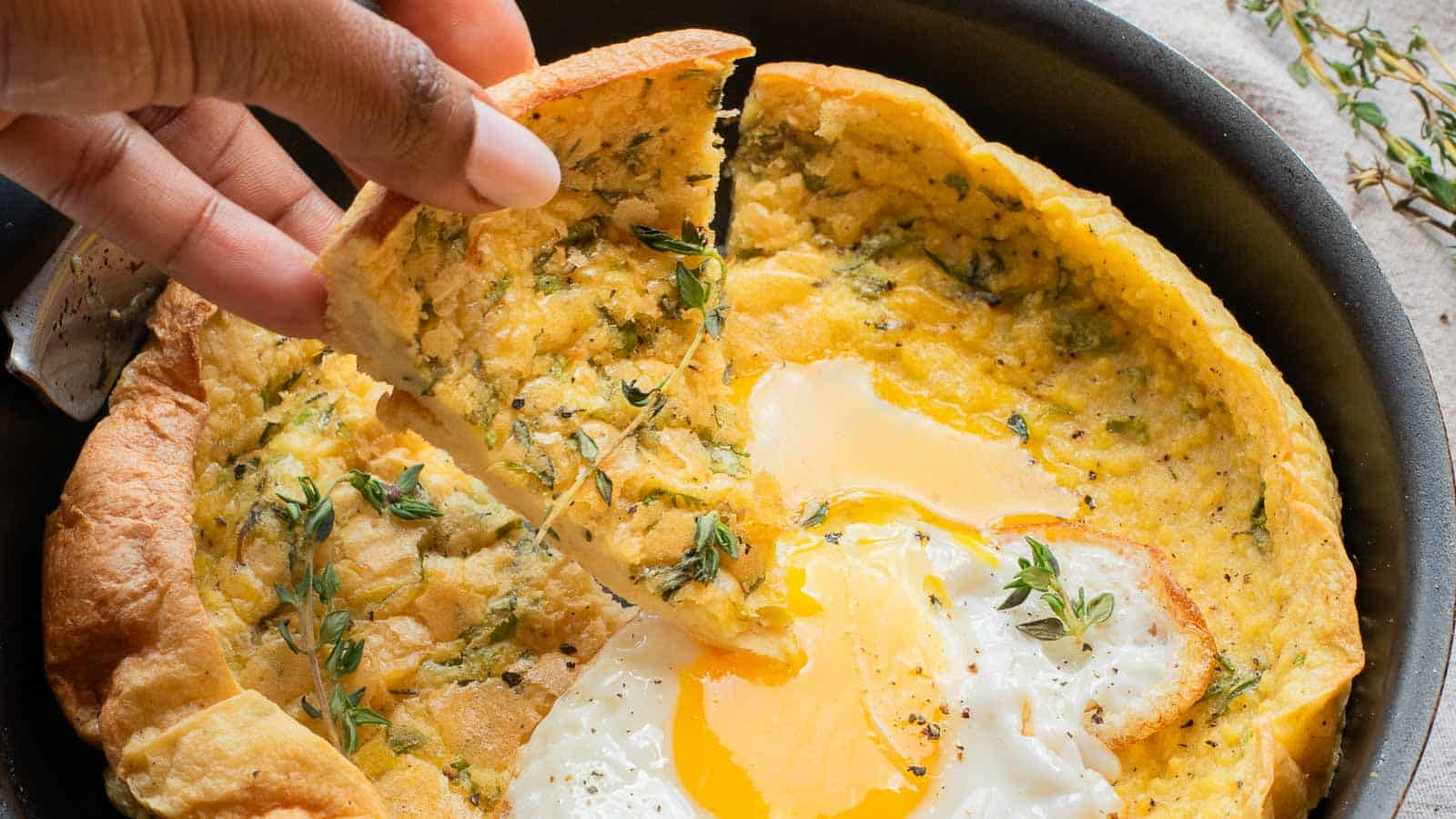 A hand lifting a slice of savory Dutch Baby pancake, crowned with a perfectly cooked fried egg and garnished with fresh herbs, in a skillet.