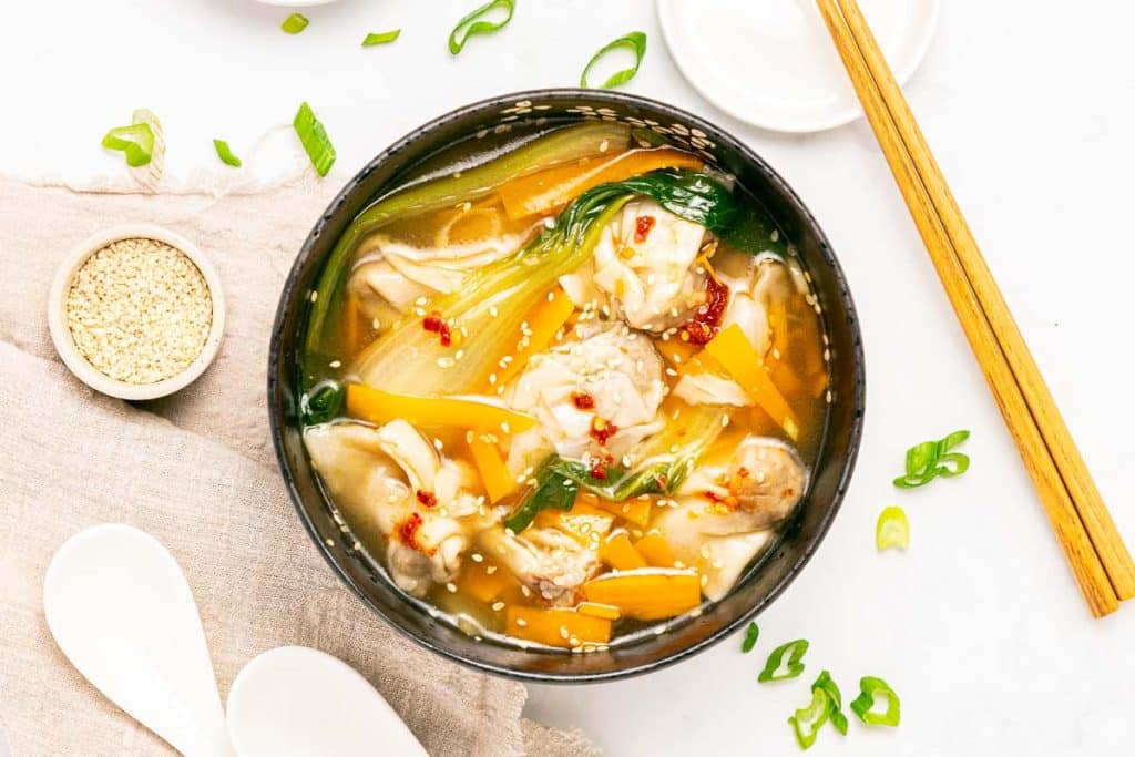 A bowl of Asian-style soup with vegetables, dumplings, and sesame seeds, garnished with green onions. Chopsticks, a small bowl of sesame seeds, and white spoons are placed beside the bowl on a light surface.