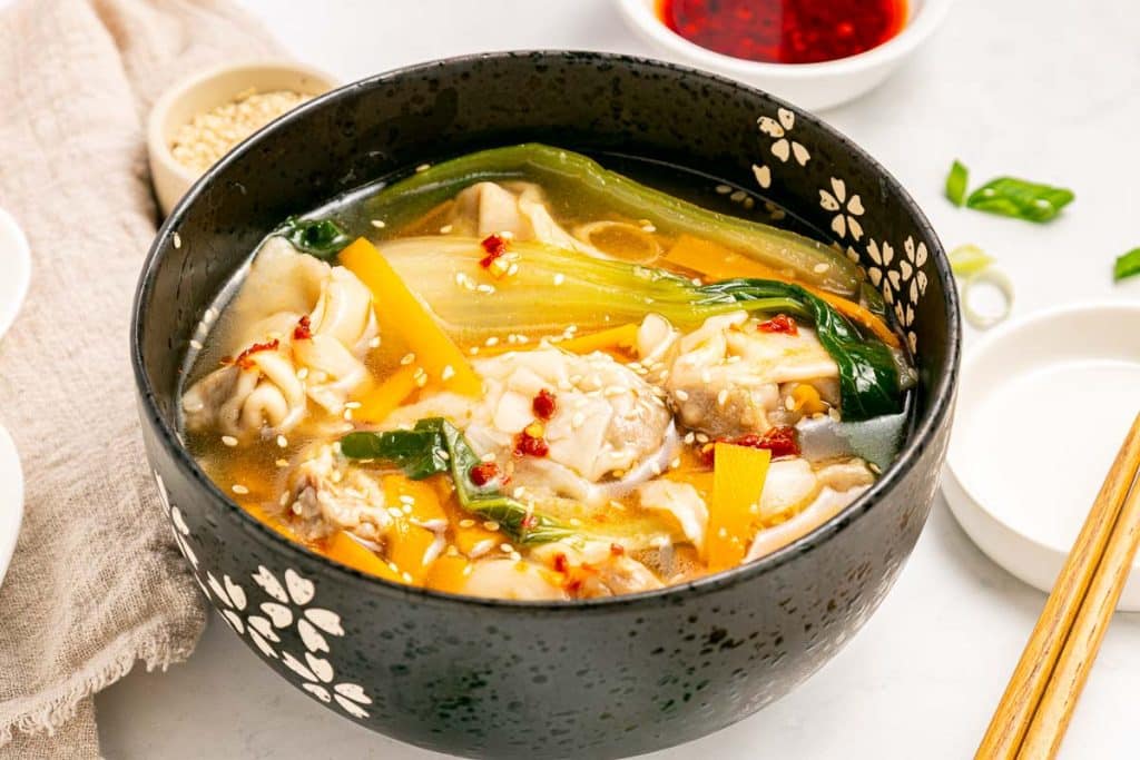 A black bowl with a floral pattern filled with wonton soup, featuring wontons, greens, yellow vegetables, and chili oil on top. Chopsticks, sauce dishes, and sesame seeds are nearby on a light surface.