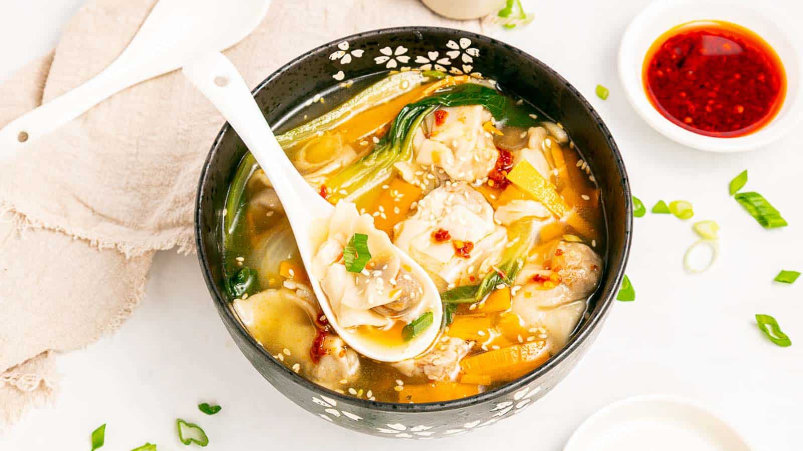 A bowl of wonton soup with dumplings, leafy greens, and sliced green onions in clear broth. A spoon holds a dumpling above the bowl. Nearby are chili oil, a white spoon, and a napkin on a white surface.