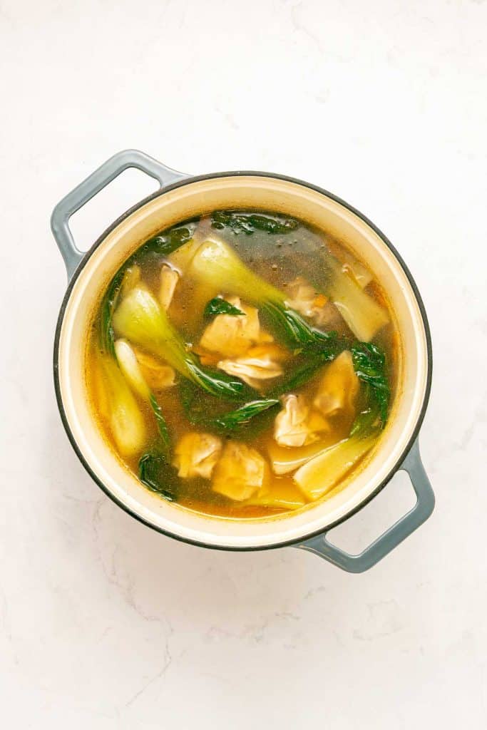A pot of soup with dumplings and leafy greens, likely bok choy, in a clear broth, sitting on a light-colored surface.