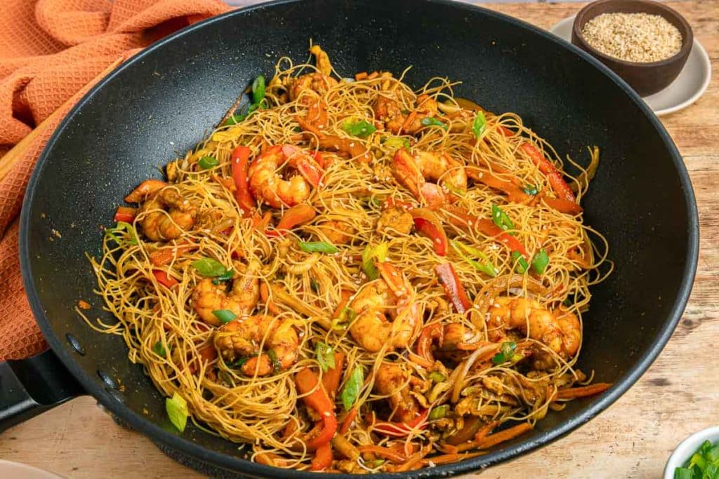 A wok filled with stir-fried rice noodles, shrimp, sliced bell peppers, green onions, and vegetables sits on a wooden table next to a bowl of sesame seeds and an orange textured cloth.