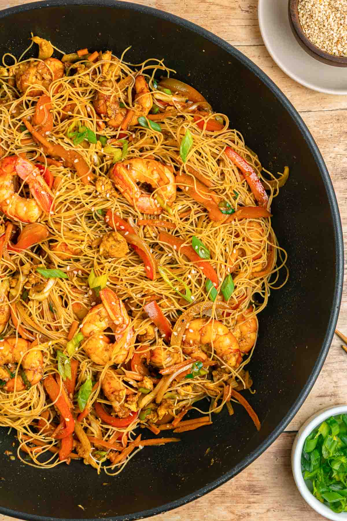 A black wok filled with stir-fried rice noodles, shrimp, red bell peppers, and green onions, garnished with sesame seeds. Small bowls of green onions and sesame seeds sit nearby on a wooden surface.