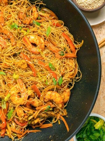 A wok filled with stir-fried rice noodles, shrimp, sliced red bell peppers, onions, and green onions, garnished with sesame seeds. Small bowls of sesame seeds and chopped green onions are beside the wok.